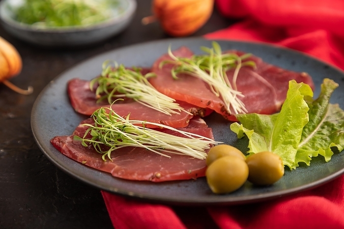 Slices of smoked salted meat with cilantro microgreen on black concrete background and red textile. Side view, close up, selective focus, by ULADZIMIR ZGURSKI