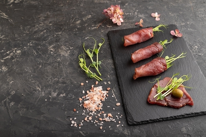 Slices of smoked salted meat with green pea microgreen on black concrete background. Side view, copy space, by ULADZIMIR ZGURSKI