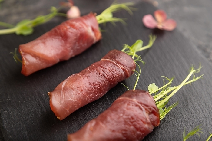 Slices of smoked salted meat with green pea microgreen on black concrete background. Side view, close up, selective focus, by ULADZIMIR ZGURSKI