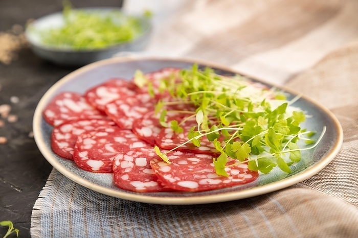 Slices of smoked cervelat salami sausage with spinach microgreen, salt and pepper on black concrete background and beige textile. Side view, close up, selective focus, by ULADZIMIR ZGURSKI