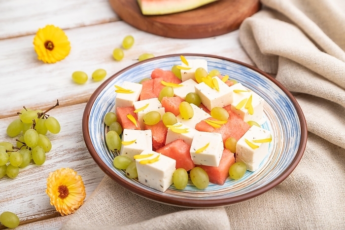 Vegetarian salad with watermelon, feta cheese, and grapes on blue ceramic plate on white wooden background and linen textile. Side view, close up, selective focus, by ULADZIMIR ZGURSKI