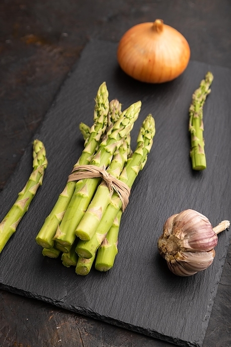 Bunch of fresh green asparagus, garlic, onion on slate board on black concrete background. Side view, close up. harvest, healthy, vegan food, concept, by ULADZIMIR ZGURSKI