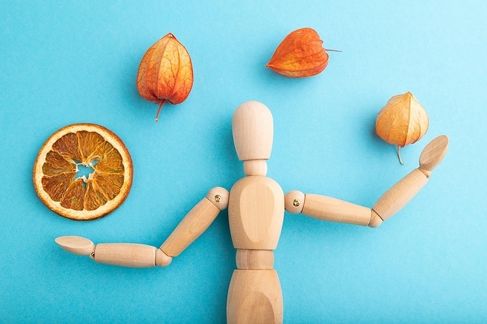 Wooden mannequin juggling dried citrus and physalis on blue pastel background. close up, isolated, by ULADZIMIR ZGURSKI