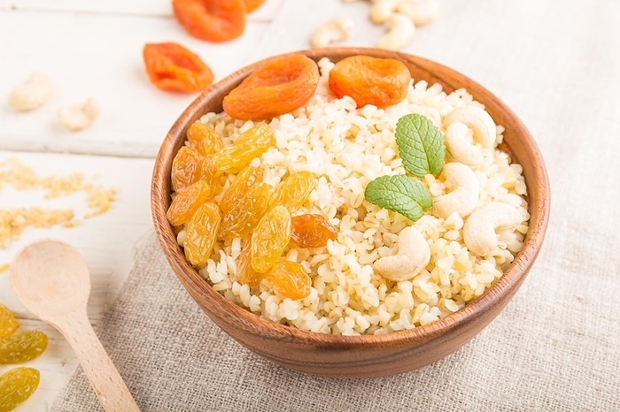Bulgur porridge with dried apricots, raisins and cashew in wooden bowl on a white wooden background and linen textile. Side view, close up, selective focus. Turkish traditional cuisine, by ULADZIMIR ZGURSKI