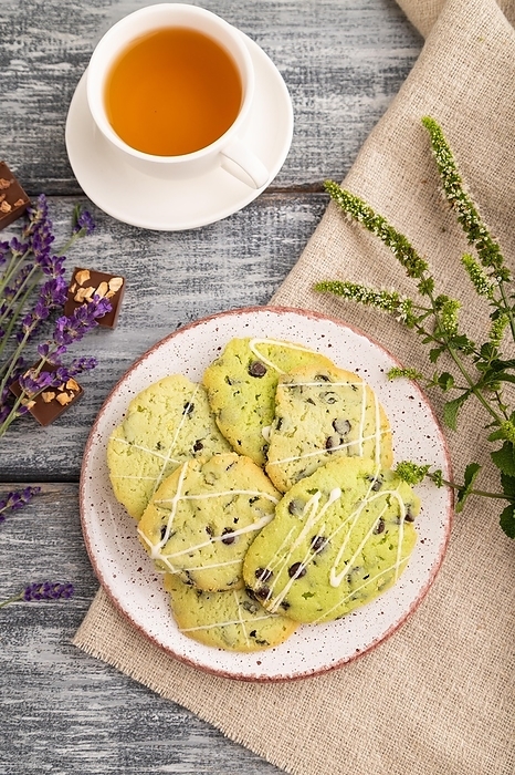 Green cookies with chocolate and mint on ceramic plate with cup of green tea and linen textile on gray wooden background. top view, flat lay, close up, by ULADZIMIR ZGURSKI