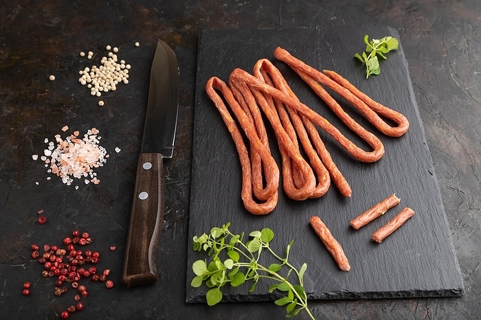 Traditional polish smoked pork sausage kabanos on a slate cutting board with salt and pepper on black concrete background. Side view, close up, by ULADZIMIR ZGURSKI