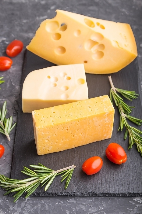 Various types of cheese with rosemary and tomatoes on black slate board on a black concrete background. Side view, close up, selective focus, by ULADZIMIR ZGURSKI