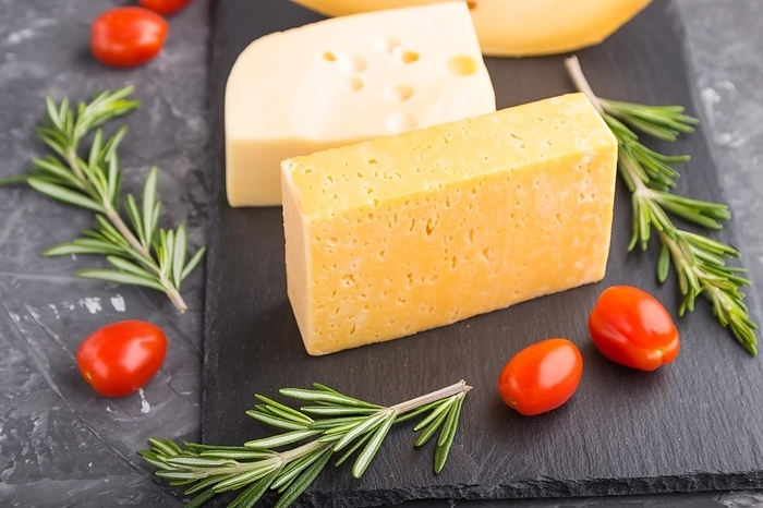 Various types of cheese with rosemary and tomatoes on black slate board on a black concrete background. Side view, close up, selective focus, by ULADZIMIR ZGURSKI