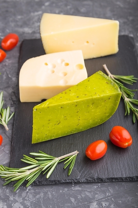 Green basil cheese and various types of cheese with rosemary and tomatoes on black slate board on a black concrete background. Side view, close up, selective focus, by ULADZIMIR ZGURSKI