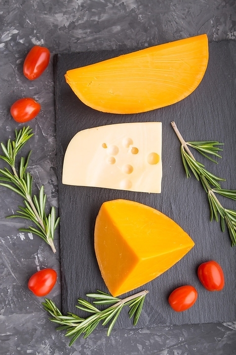Various types of cheese with rosemary and tomatoes on black slate board on a black concrete background. Top view, flat lay, close up, by ULADZIMIR ZGURSKI