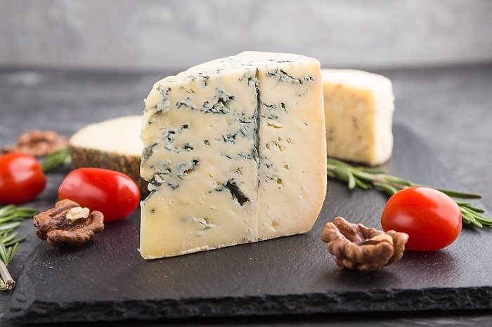 Blue cheese and various types of cheese with rosemary and tomatoes on black slate board on a black concrete background. Side view, close up, selective focus, by ULADZIMIR ZGURSKI
