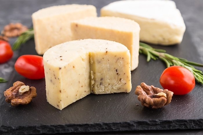 Cheddar cheese and various types of cheese with rosemary and tomatoes on black slate board on a black concrete background. Side view, close up, selective focus, by ULADZIMIR ZGURSKI