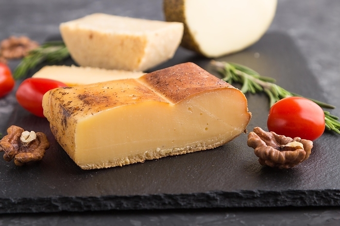 Smoked cheese and various types of cheese with rosemary and tomatoes on black slate board on a black concrete background. Side view, close up, selective focus, by ULADZIMIR ZGURSKI