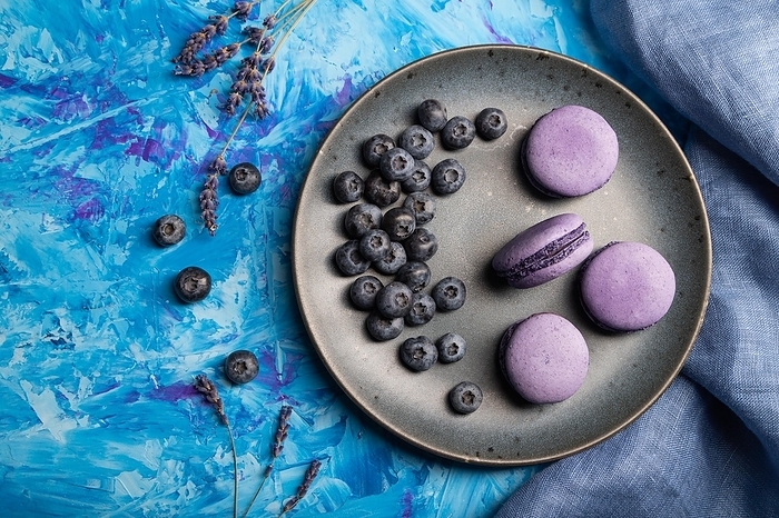 Purple macarons or macaroons cakes with blueberries on ceramic plate on a blue concrete background and blue textile. Flat lay, top view, close up, by ULADZIMIR ZGURSKI