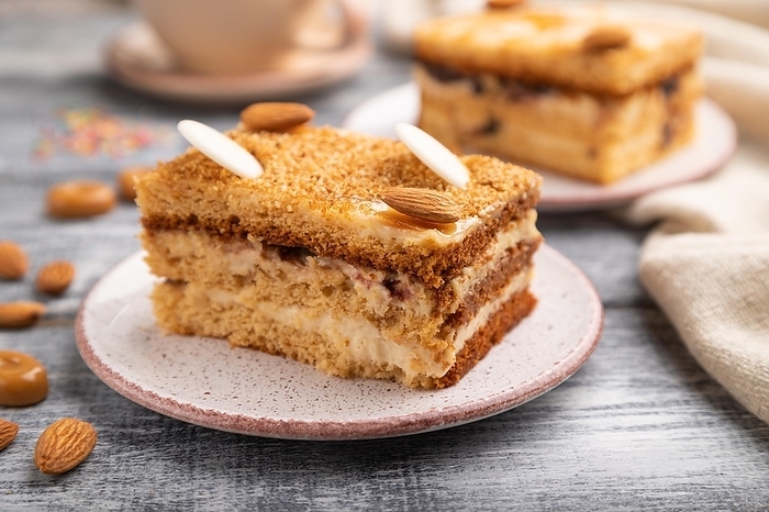 Honey cake with milk cream, caramel, almonds and a cup of coffee on a gray wooden background and linen textile. Side view, close up, selective focus, by ULADZIMIR ZGURSKI