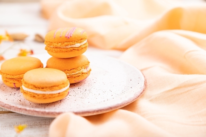Orange macarons or macaroons cakes with cup of apricot juice on a white wooden background and orange linen textile. Side view, close up, selective focus, by ULADZIMIR ZGURSKI