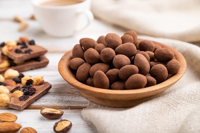 Almond in chocolate dragees in wooden plate and a cup of coffee on white wooden background and linen textile. Side view, close up, selective focus, by ULADZIMIR ZGURSKI