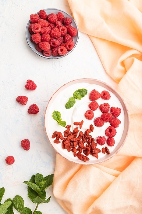 Yogurt with raspberry and goji berries in ceramic bowl on white concrete background and orange linen textile. top view, flat lay, close up, by ULADZIMIR ZGURSKI