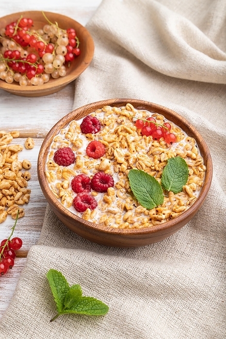 Wheat flakes porridge with milk, raspberry and currant in wooden bowl on white wooden background and linen textile. Side view, close up, by ULADZIMIR ZGURSKI