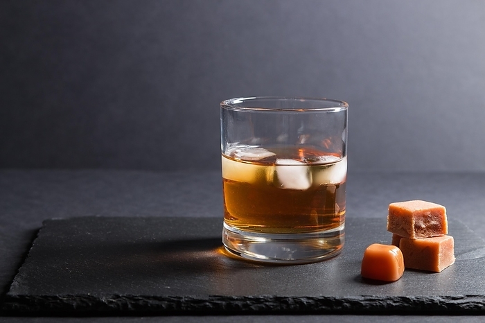 Glass of amber whiskey with ice and caramel candies on a black stone slate board on black background. Side view, close up, selective focus, copy space, by ULADZIMIR ZGURSKI