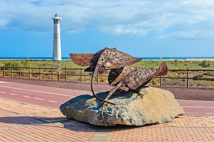 In the foreground sculpture made of scrap metal of marine animals two stingrays rays on stone pedestal, behind it developed cycle path wide cycle road with markings, left in the background lighthouse Faro de Morro Jable, Morro Jable, Jandia peninsula, Fuerteventura, Canary Islands, Canary Islands, Spain, Europe, by Frank Schneider