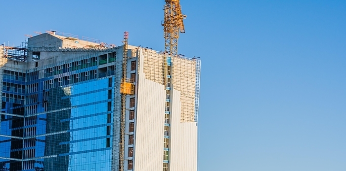 Yellow crane elevator attached to side of building that is under construction against a clear blue sky, by John Erskin