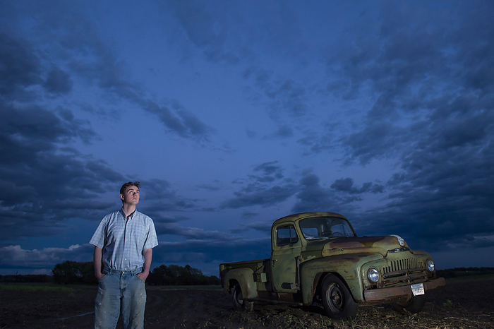JOSAR_sartore_cole Young man looks at the gathering storm clouds in the sky near a 1951 International Harvester pickup truck on a farm field  Bennet, Nebraska, United States of America, by Joel Sartore Photography   Design Pics
