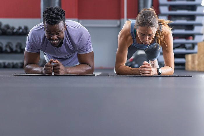 Fit diverse couple doing planks at the gym. Their workout routine emphasizes core strength and partner motivation.