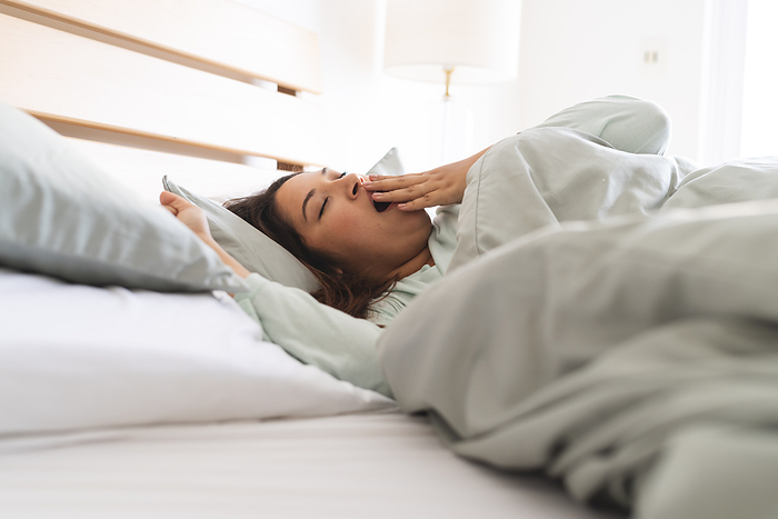Young plus size Biracial woman waking up in a cozy bedroom, with copy space unaltered. She's checking her phone and yawning, starting her day at home.
