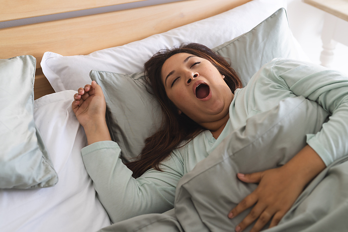 Young biracial plus size woman waking up in a cozy bedroom, with copy space unaltered. She's captured mid-yawn, suggesting a fresh start to her day at home.