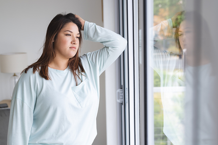 Young plus size biracial woman looks contemplative by a window at home. She appears reflective, gazing outside, lost in thought in a serene indoor setting unaltered.