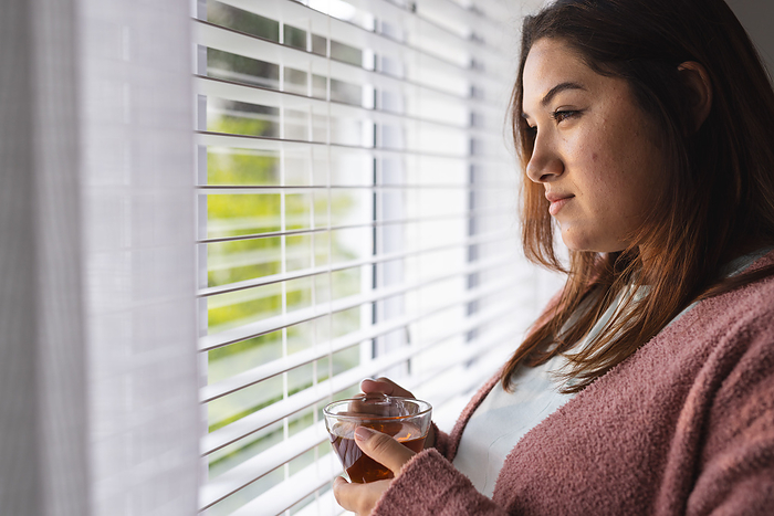 Young plus size biracial woman gazes outside a window, holding a warm beverage. She appears contemplative, unaltered, enjoying a quiet moment at home.