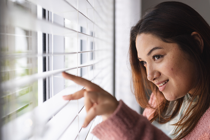 Young biracial plus size woman peers through blinds at home, with copy space. Her curious gaze adds a sense of anticipation or privacy to the scene, unaltered.