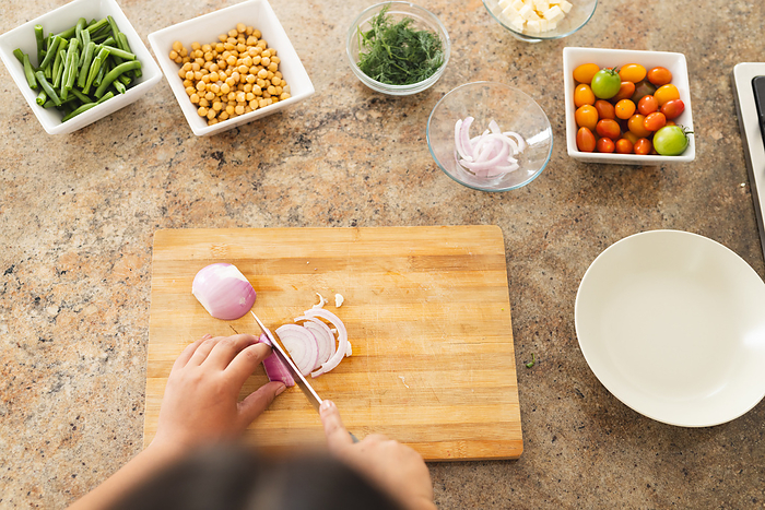 Hands of a person slicing onion on a wooden cutting board in a home kitchen. Fresh ingredients await to be combined into a nutritious meal, emphasizing healthy eating.