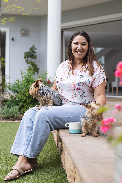 Young plus size biracial woman enjoys a relaxed moment outdoors with her dogs. She's seated on a step at home, embodying a casual lifestyle with her pets unaltered.
