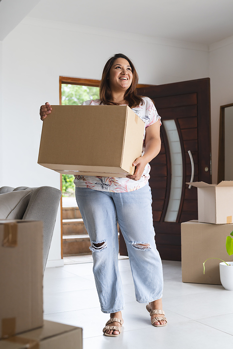 Young biracial plus size woman carries a moving box at home, with copy space. She smiles brightly, suggesting excitement about a new beginning unaltered.