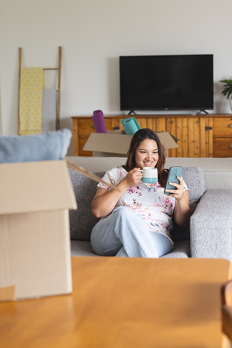 Young plus size biracial woman enjoys a break at home, surrounded by moving boxes. She sips coffee and checks her phone, settling into her new space unaltered.