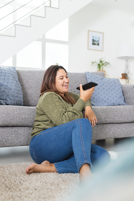 Young plus size biracial woman enjoys leisure time at home, with copy space unaltered. Sitting on the floor, she's engaged with her smartphone, exuding a relaxed vibe.
