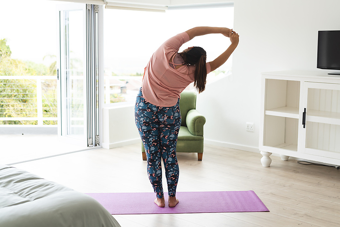 Young plus size biracial woman stretches on a yoga mat at home, with copy space unaltered. She practices morning wellness routine in a bright, airy living space.