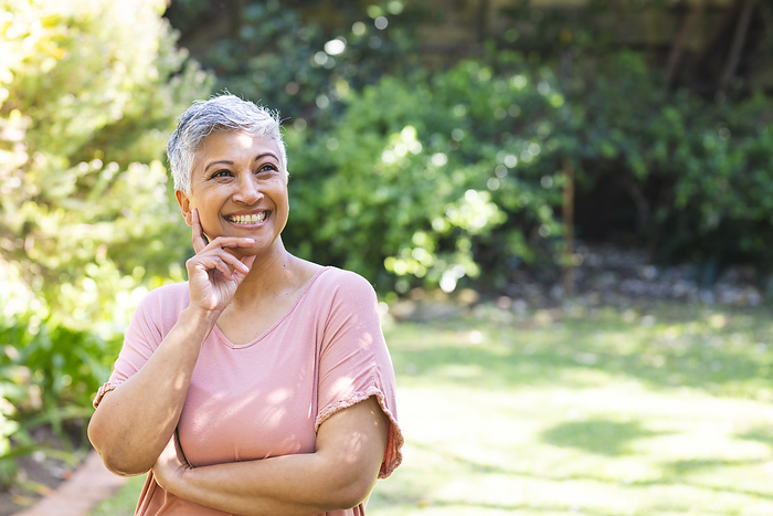 A mature biracial senior woman enjoys a sunny day in the garden, with copy space unaltered. Her cheerful expression reflects the tranquility of a relaxing outdoor setting.