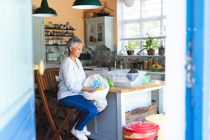 A mature biracial woman sorts recycling at home, with copy space. She contributes to environmental sustainability from her kitchen unaltered.