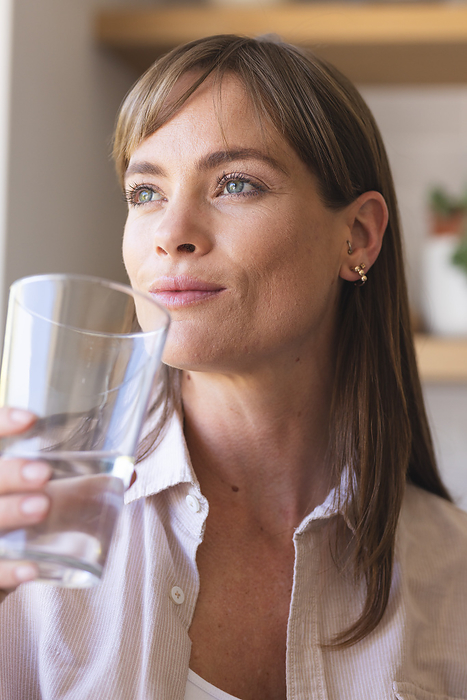 A middle-aged Caucasian woman is drinking water from a clear glass. Her gaze appears thoughtful and unaltered, emphasizing the importance of hydration for health and well-being.