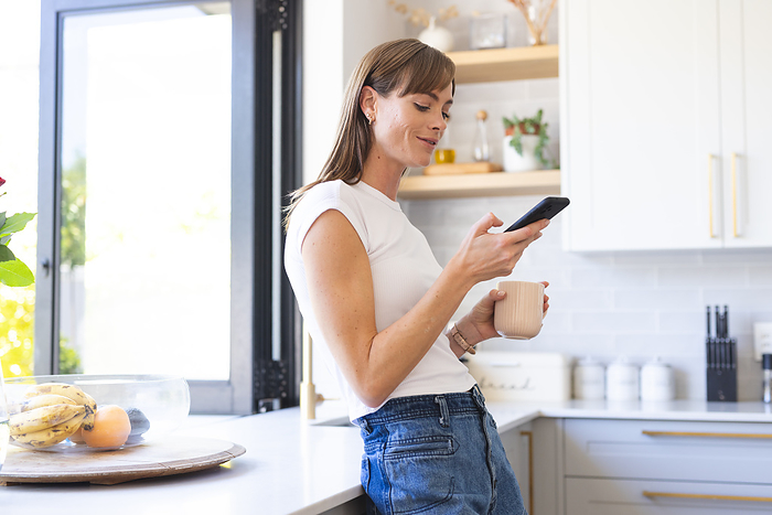 A young Caucasian woman is casually using her smartphone in a bright kitchen. She appears relaxed, browsing social media or texting as she enjoys a cup of coffee unaltered.
