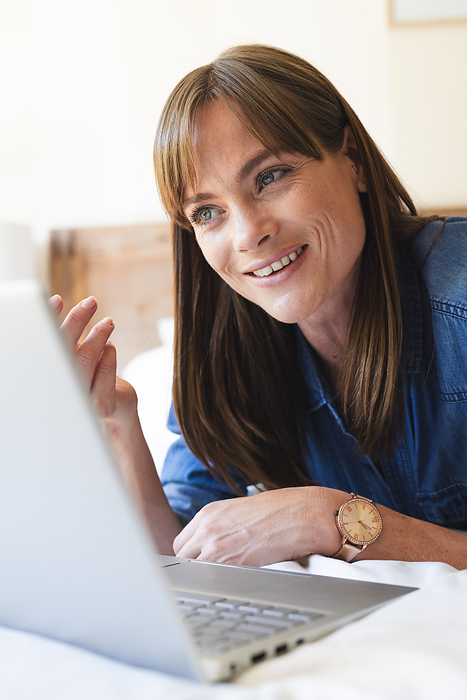 A middle-aged Caucasian woman smiles while engaging in a video call on her laptop. Her friendly demeanor suggests she's having a pleasant conversation or virtual meeting unaltered.