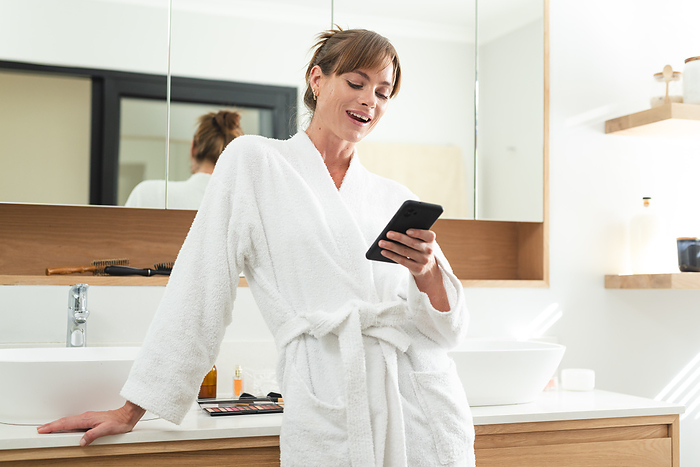 Caucasian middle-aged woman in a white robe enjoys her phone at home. Her cheerful demeanor suggests a positive interaction or delightful news.