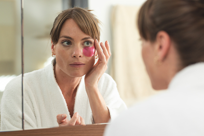 Caucasian woman examines her face in the mirror, applying a pink eye mask. She appears concerned about her skin condition at home.