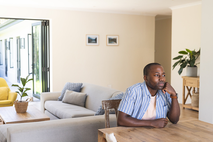 Young African American man seated thoughtfully at home, with copy space. He appears contemplative in a well-lit, cozy living room setting.