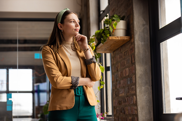 Young Caucasian woman stands thoughtfully by the office window in a casual business setting. She appears contemplative in a stylish environment.