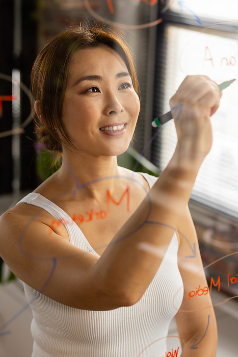 Asian woman writes on a glass board in a casual business office setting. Her focus suggests strategic planning or creative brainstorming at work.