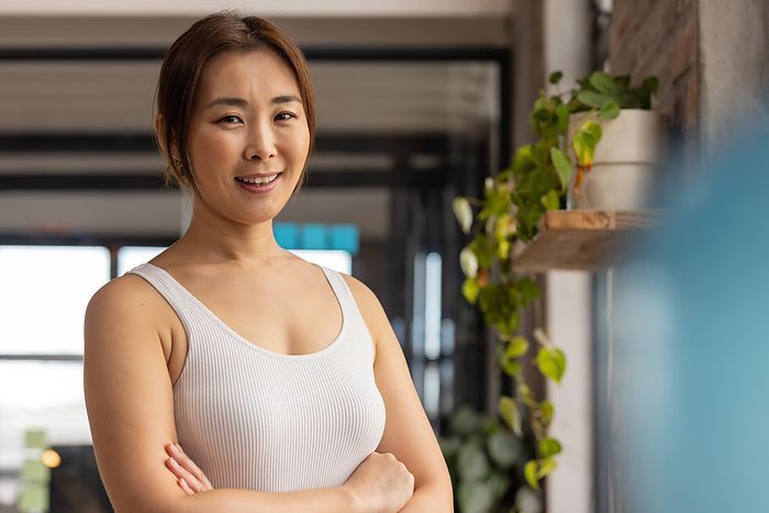 A confident Asian woman stands in a modern, casual business office, with copy space. Her arms are crossed, exuding professionalism and poise in the workplace.